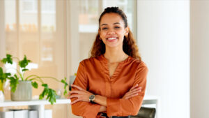 confident stylish woman laughing with arms crossed and standing inside a modern office near a window. face portrait of a happy hispanic female entrepreneur looking cheerful about successful business